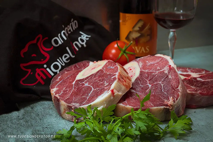 fotografía de un bodegón con carne de ossobuco, una botella de vino, una copa y un tomate
